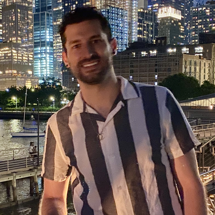 Photo of Gay For Good NYC chapter leader, Steve Mortenson (he/him), standing along side water with tall buildings lit up behind him.
