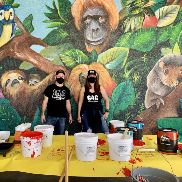 Two volunteers standing in front of a mural of animals with a table and paint supplies in front of them