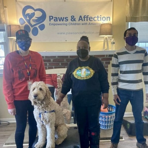 Two women and a man with a large white dog standing side by side in front of a sign that says 'Paws & Affection'