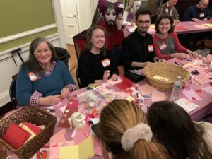 Group of smiling volunteers at a table strewn with Valentine's themed card making supplies. 